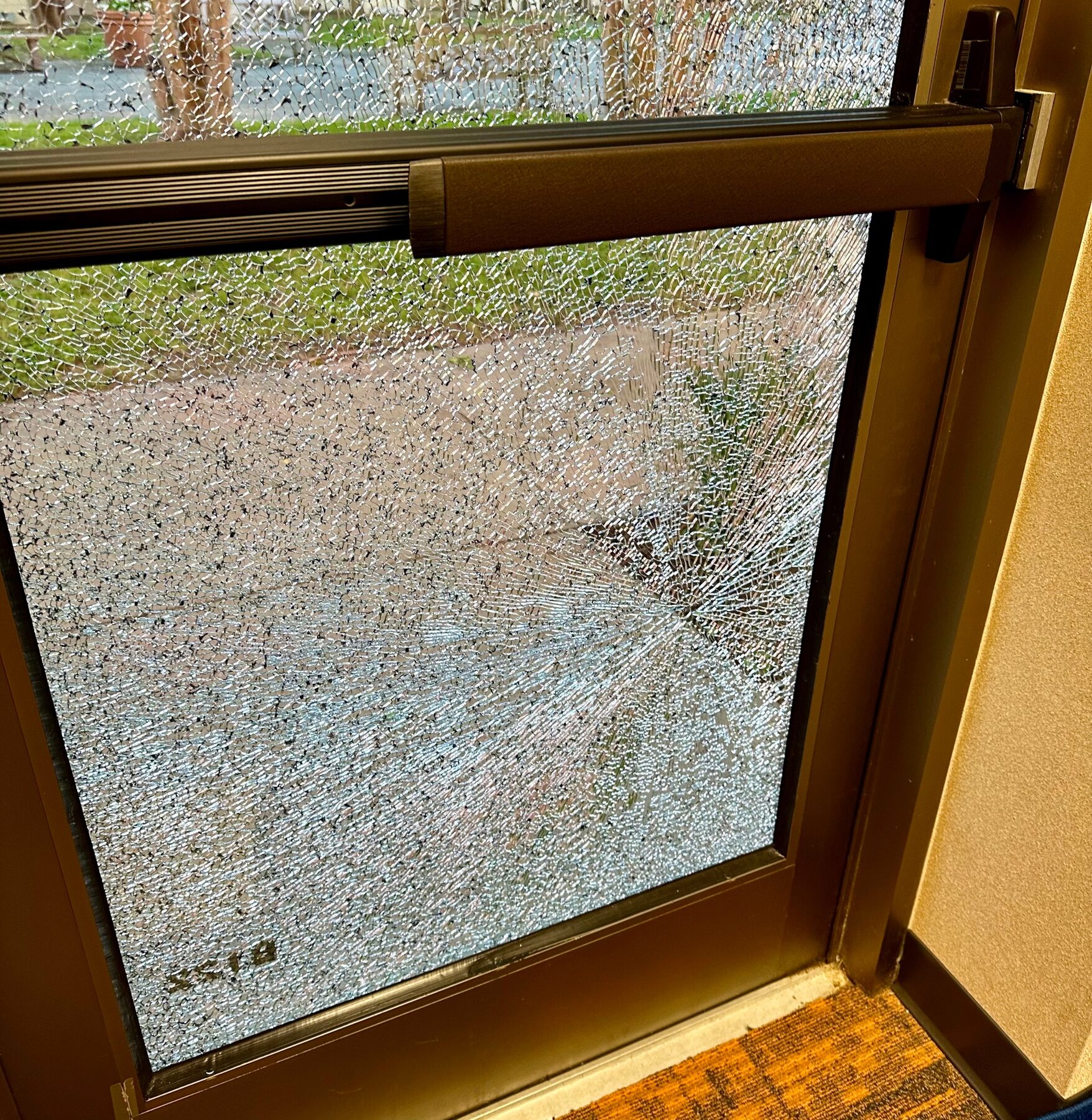 School exit door with a panic bar, tempered glass shattered across the whole panel by a thrown rock but held intact in its frame by 8 mil security film